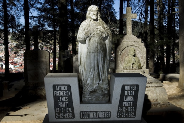 A stone statue of Jesus stands amidst old gravestones in a cemetery surrounded by trees. Two prominent gravestones in front read Father Frederick James Pratt and Mother Hilda Laura Pratt with the inscription 'Together Forever.' In the background, there is a view of a city partially obscured by shadows from the trees.