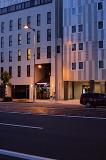 A nighttime urban street scene with a multistory building featuring a hotel entrance. Soft streetlights cast a warm glow on the sidewalk, highlighting a cyclist passing by. A red car is parked on the side of the road, and the building exterior is a mix of white and gray panels with several dark windows.