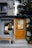 A quaint storefront with a wooden door and a large window displaying various items inside. The exterior is partially covered with green ivy, adding an element of nature. A small bench and a potted plant sit in front of the entrance. Above the door, various posters and flyers are visible.