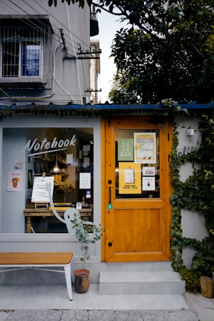 A quaint storefront with a wooden door and a large window displaying various items inside. The exterior is partially covered with green ivy, adding an element of nature. A small bench and a potted plant sit in front of the entrance. Above the door, various posters and flyers are visible.