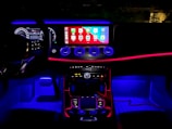 A car interior with ambient lighting, featuring a sleek dashboard. The central display shows various app icons, illuminated by vibrant blue and red lighting. The steering wheel's digital display shows driving information, while three circular air vents stand out below the screen.