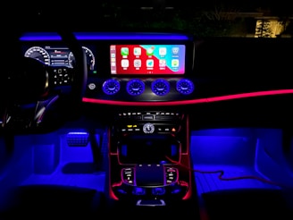 A car interior with ambient lighting, featuring a sleek dashboard. The central display shows various app icons, illuminated by vibrant blue and red lighting. The steering wheel's digital display shows driving information, while three circular air vents stand out below the screen.