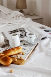 A cozy breakfast scene on a bed with a white setting. Two baguettes are placed on a wooden tray alongside a few scattered nuts. A white teacup with a saucer and a white milk jug sit nearby. There's an open magazine adding to the relaxed atmosphere. Soft, neutral tones create a serene and calming feeling.