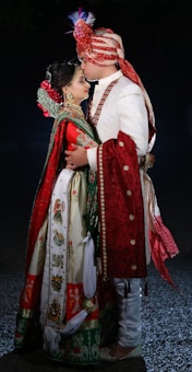 A couple is dressed in traditional Indian wedding attire, with intricate embroidery and vivid colors. The man wears a turban and a white sherwani with red accents, while the woman is adorned in a colorful lehenga with heavy jewelry and a floral hairpiece. They are embracing closely.