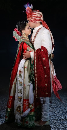 A couple is dressed in traditional Indian wedding attire, with intricate embroidery and vivid colors. The man wears a turban and a white sherwani with red accents, while the woman is adorned in a colorful lehenga with heavy jewelry and a floral hairpiece. They are embracing closely.