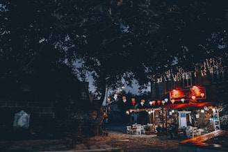 A cozy outdoor cafe set in a dimly lit street with string lights and colorful paper lanterns hanging above. The stone building features warm lighting, creating a welcoming ambiance. Tables and chairs are arranged beneath the canopy of an old tree next to a stone wall, adding a rustic charm.