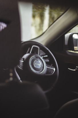 A close-up view of a car's interior, focusing on the steering wheel with a Jeep logo. The dashboard and a part of the side mirror are visible, creating a cozy, enclosed automotive atmosphere.