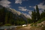 A camping scene set in a mountainous region with tall pine trees under a starry night sky. A white car is parked beside a pitched tent near a flowing stream. The mountains are illuminated, possibly by moonlight or early evening light, adding depth to the landscape.