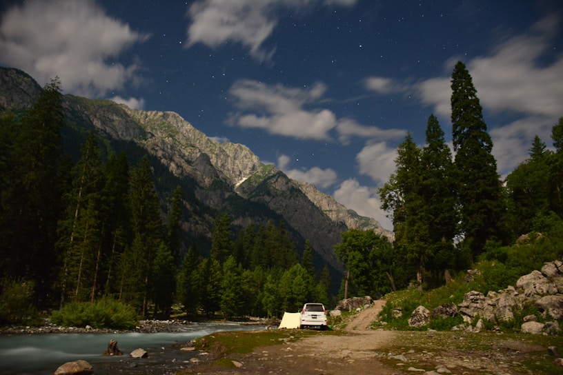 A camping scene set in a mountainous region with tall pine trees under a starry night sky. A white car is parked beside a pitched tent near a flowing stream. The mountains are illuminated, possibly by moonlight or early evening light, adding depth to the landscape.