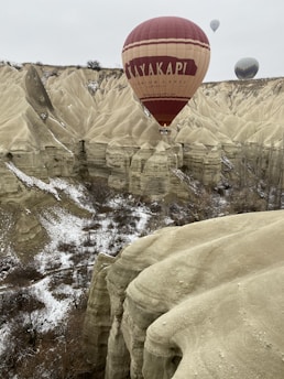A hot air balloon with the words 'Kayakapi Premium Caves Cappadocia' is floating over a rugged, sandy landscape with unique rock formations dusted with snow. The terrain is characterized by steep, eroded cliffs and scattered vegetation. In the background, additional hot air balloons are visible against the overcast sky.