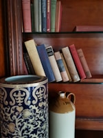 Close-up of a modern ceramic vase in soft brown resting on a light wooden shelf among books.