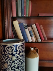 An antique vase with intricate patterns sitting on a vintage shelf surrounded by old books.