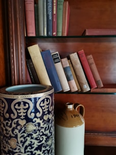 An antique vase with intricate patterns sitting on a vintage shelf surrounded by old books.