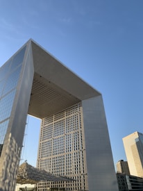 A large, modern arch-shaped building with a grid-like facade stands against a clear blue sky. The structure is characterized by its angular design and reflective glass surfaces. Nearby, other contemporary skyscrapers are visible, contributing to an urban landscape.
