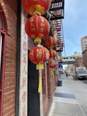 A series of red paper lanterns with gold Chinese characters hang against a brick building. The scene is set in an urban environment with city infrastructure visible in the background, including fire escapes and a busy street. The entrance is framed by red and beige columns with more Chinese characters inscribed.
