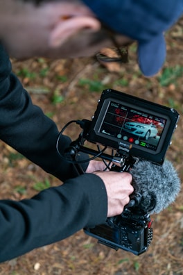 A person wearing a navy-blue cap and black long-sleeved shirt is operating a camera with a mounted monitor. The monitor displays an image of a white car. The person appears to be outdoors, as the ground is covered with brown pine needles and green patches of grass.