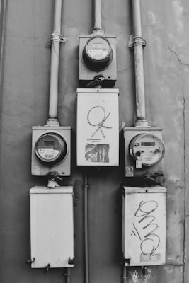 An array of electrical meters mounted on a wall with visible pipes and wires. The meters are housed in rectangular metal boxes with some graffiti on their surfaces. The overall appearance is industrial and functional with signs of wear.