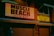 A dimly lit urban scene featuring bright neon signs reading 'Muscle Beach Juice Bar' and 'Nutrition' on the façade of a building. The setting appears to be evening or night, with an atmosphere created by the glowing signage. A person wearing a mask is visible in the foreground, suggesting a pandemic era.
