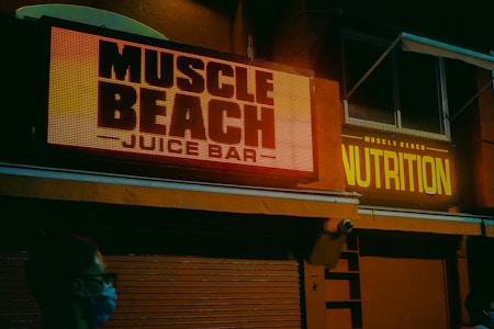 A dimly lit urban scene featuring bright neon signs reading 'Muscle Beach Juice Bar' and 'Nutrition' on the façade of a building. The setting appears to be evening or night, with an atmosphere created by the glowing signage. A person wearing a mask is visible in the foreground, suggesting a pandemic era.