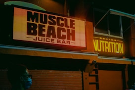 A dimly lit urban scene featuring bright neon signs reading 'Muscle Beach Juice Bar' and 'Nutrition' on the façade of a building. The setting appears to be evening or night, with an atmosphere created by the glowing signage. A person wearing a mask is visible in the foreground, suggesting a pandemic era.