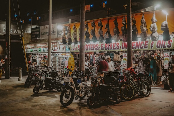 A bustling street market at night with bright lights highlighting various items for sale. Scooters and e-bikes are parked in the foreground, while several people browse through hanging bags and clothing beneath a brightly lit awning with signage for items like incense and oils. The market has an eclectic and vibrant atmosphere.