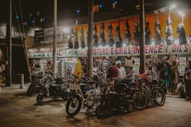 A bustling street market at night with bright lights highlighting various items for sale. Scooters and e-bikes are parked in the foreground, while several people browse through hanging bags and clothing beneath a brightly lit awning with signage for items like incense and oils. The market has an eclectic and vibrant atmosphere.