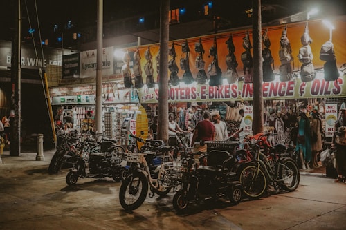 A bustling street market at night with bright lights highlighting various items for sale. Scooters and e-bikes are parked in the foreground, while several people browse through hanging bags and clothing beneath a brightly lit awning with signage for items like incense and oils. The market has an eclectic and vibrant atmosphere.