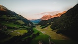A picturesque landscape featuring a lush green valley nestled between rolling hills. Small houses and farms dot the landscape, with winding roads leading through the valley. In the background, majestic mountains are illuminated by the warm glow of a sunset.