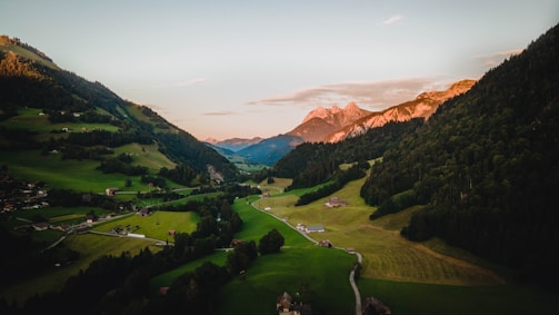 A picturesque landscape featuring a lush green valley nestled between rolling hills. Small houses and farms dot the landscape, with winding roads leading through the valley. In the background, majestic mountains are illuminated by the warm glow of a sunset.
