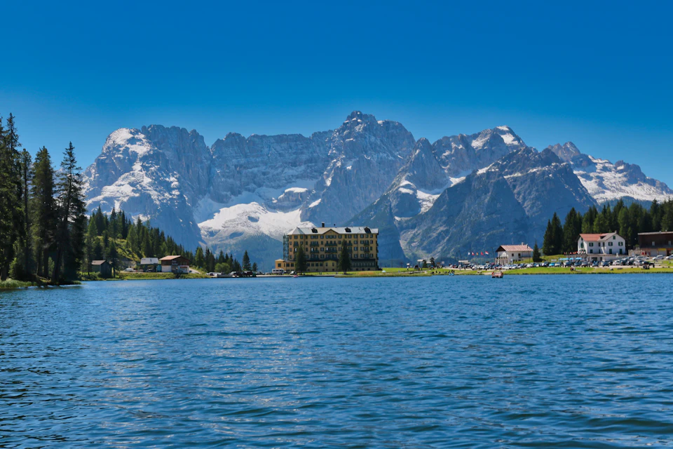 The image depicts a serene mountain lake surrounded by snow-capped mountains and lush green trees. The calm, blue waters of the lake reflect the clear sky above. In the distance, there is a large building with a yellow roof nestled among the mountains. Several boats are docked at the shoreline, suggesting that this location may be a popular spot for boating or other water-based activities.