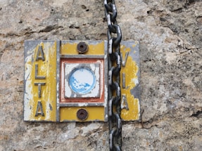 A weathered and worn sign attached to a rough stone surface, featuring the word 'ALTA' on one side and 'VIA' on the other. The sign has chipped yellow paint, a rusted circular metal detail, and a thick black chain draped across it.