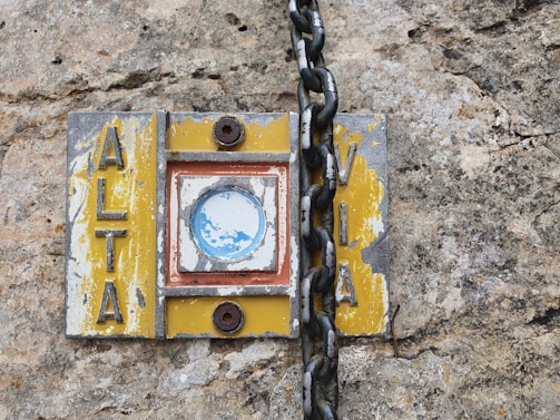 A weathered and worn sign attached to a rough stone surface, featuring the word 'ALTA' on one side and 'VIA' on the other. The sign has chipped yellow paint, a rusted circular metal detail, and a thick black chain draped across it.