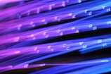 Brightly colored lines of purple and blue light create an abstract pattern, with scattered letters or symbols appearing intermittently throughout the image. The effect resembles light trails or fiber optic cables in motion.