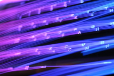 Brightly colored lines of purple and blue light create an abstract pattern, with scattered letters or symbols appearing intermittently throughout the image. The effect resembles light trails or fiber optic cables in motion.