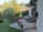A quaint patio area outside a house featuring stone flooring and surrounded by lush greenery. There is a wooden bench with a potted plant nearby and a table covered with a cloth, accompanied by folding chairs. Flower pots with colorful blossoms enhance the charming outdoor setting.