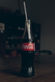 A classic glass bottle of Coca-Cola is placed upright on a wooden table. The bottle contains a dark, carbonated liquid and has a red label with white text. A straw is inserted into the bottle. The background is dimly lit, providing a contrast to the bottle.
