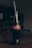 A classic glass bottle of Coca-Cola is placed upright on a wooden table. The bottle contains a dark, carbonated liquid and has a red label with white text. A straw is inserted into the bottle. The background is dimly lit, providing a contrast to the bottle.