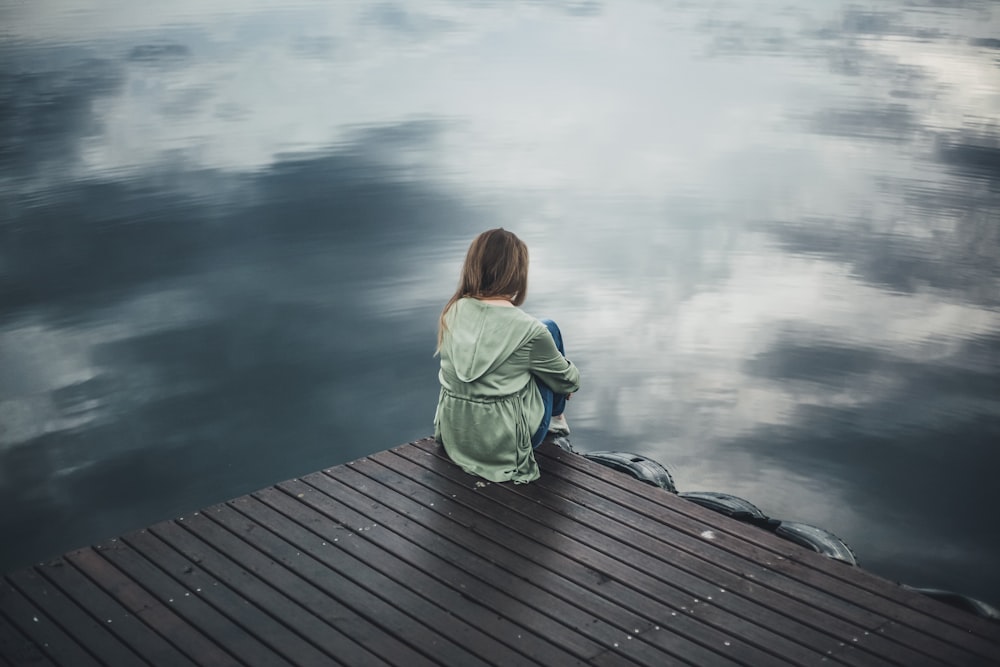 woman in green hoodie sitting on dock looking at body of water