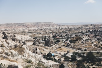 A panoramic view of a unique landscape featuring eroded rock formations and a small settlement nestled among them. The terrain is arid and rugged, with various conical and fairy chimney-like structures dotting the scene. Sparse vegetation is seen, primarily shrubs and small trees.