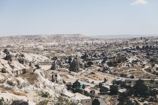 A panoramic view of a unique landscape featuring eroded rock formations and a small settlement nestled among them. The terrain is arid and rugged, with various conical and fairy chimney-like structures dotting the scene. Sparse vegetation is seen, primarily shrubs and small trees.