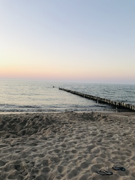 The image depicts a serene beach scene at sunset. The horizon shows a gradient sky transitioning from soft orange to light blue. The calm sea meets the sandy beach in the foreground, where a pair of flip-flops lie on the sand. A wooden breakwater extends into the ocean, creating a sense of depth.