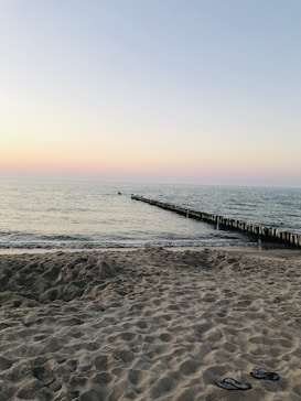 The image depicts a serene beach scene at sunset. The horizon shows a gradient sky transitioning from soft orange to light blue. The calm sea meets the sandy beach in the foreground, where a pair of flip-flops lie on the sand. A wooden breakwater extends into the ocean, creating a sense of depth.