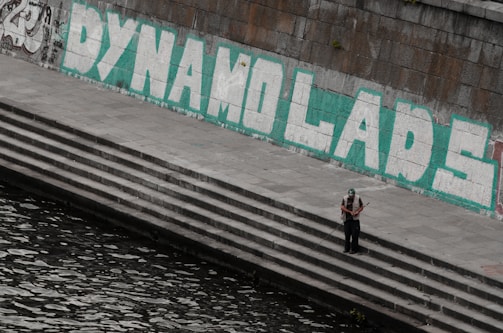A large graffiti with the words 'DYNAMO LADS' in bold white letters outlined in green is painted on a stone wall above a series of concrete steps. A person wearing a cap stands alone on the steps near the water, holding a fishing rod.