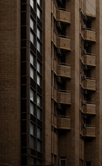 A tall building with a facade made of brown bricks. The structure features multiple small balconies, each equipped with an air conditioning unit. The windows are modern and vertically aligned, contributing to a streamlined architectural appearance.