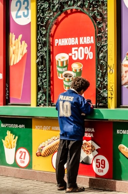 A person is standing at a fast-food stand, dressed in a blue sports jersey with the number 10 and the word 'France' on the back. The surrounding display has vibrant advertisements featuring various food items like french fries, coffee, hot dogs, and other snacks, alongside prices in Ukrainian currency. The backdrop includes bold and colorful signage, creating a lively and busy atmosphere.