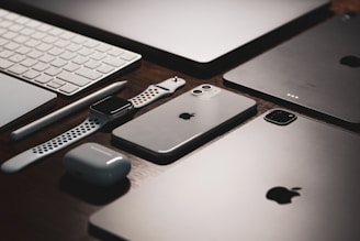 A collection of sleek electronic devices including a smartphone, smartwatch, wireless earbuds, tablet, stylus, and keyboard arranged neatly on a wooden surface.