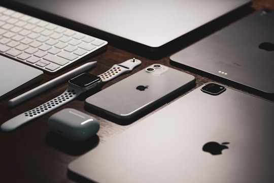 A collection of sleek electronic devices including a smartphone, smartwatch, wireless earbuds, tablet, stylus, and keyboard arranged neatly on a wooden surface.