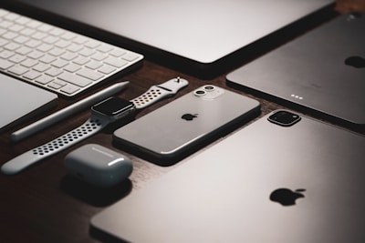 A collection of sleek electronic devices including a smartphone, smartwatch, wireless earbuds, tablet, stylus, and keyboard arranged neatly on a wooden surface.