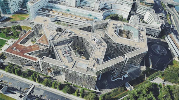 Aerial view of a large, modern architectural building with geometric shapes and a complex layout. The structure is predominantly made of glass and concrete, featuring multiple levels and intricate design elements. Surrounding the building are green lawns and other urban infrastructure.