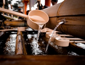 A wooden ladle is positioned under a bamboo spout with water flowing from the spout into the ladle. The arrangement is part of a traditional water purification basin, typically found at Japanese shrines. Several other ladles rest nearby, partially submerged in water.